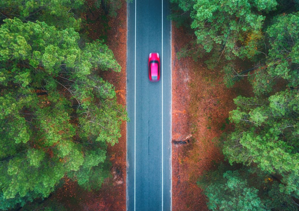 Aerial view of road with blurred car in summer forest at sunset. Amazing landscape with rural road, trees with green leaves in sunny day. Highway through the park. Top view from flying drone. Nature
