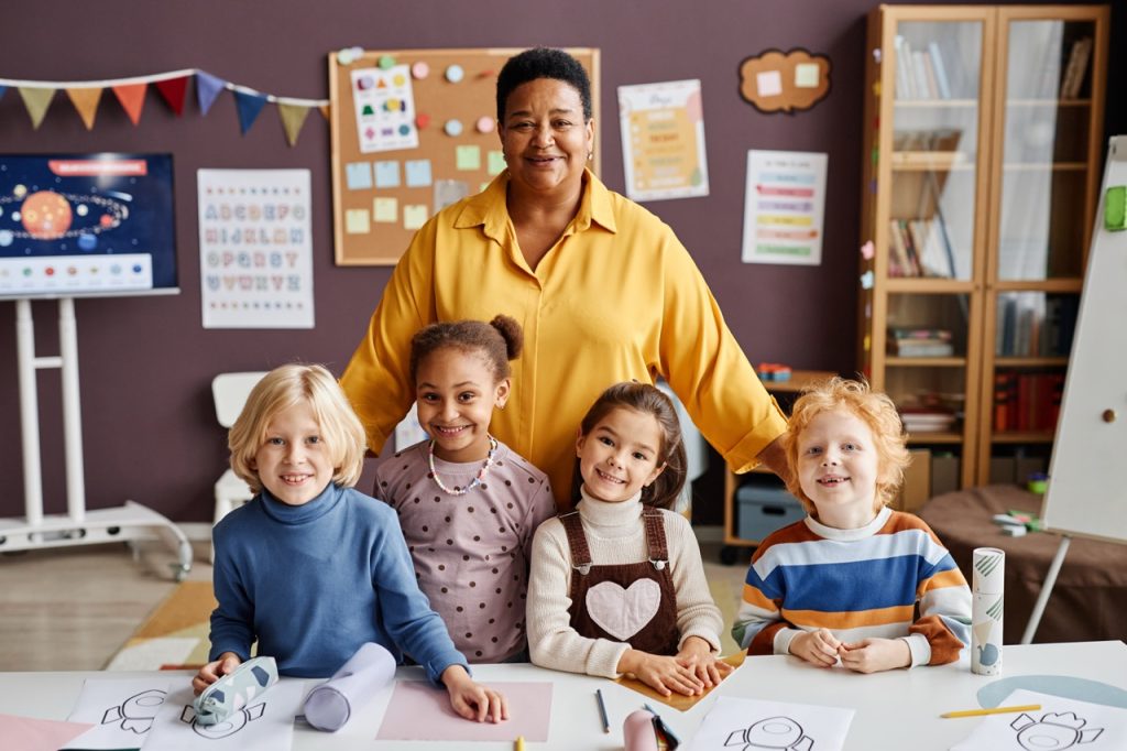 Happy mature teacher and group of intercultural little learners standing by desk and looking at camera at lesson in classroom