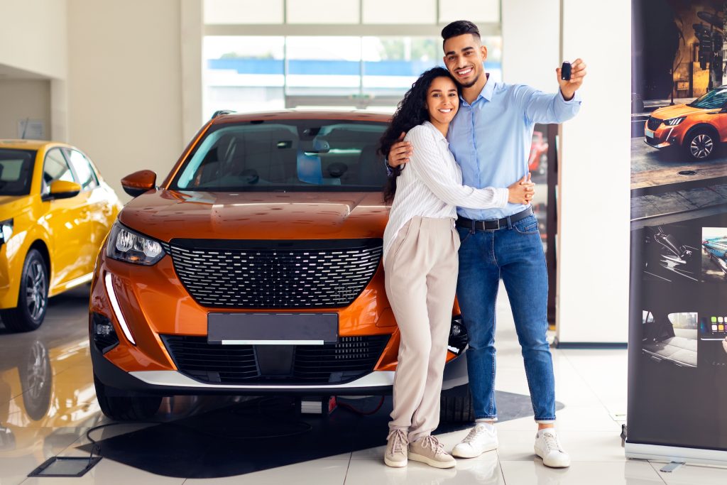 Happy middle eastern couple young man and woman standing by nice brand new car, holding key, embracing and smiling, buying new auto together at luxury showroom, panorama with copy space
