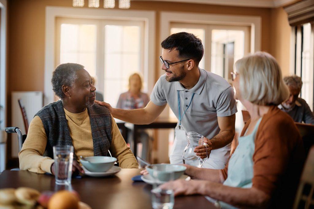 Young healthcare worker talking to seniors during lunch at nursing home.