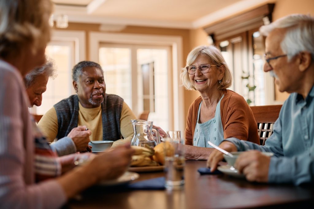 Group of senior friends communicating while eating lunch in nursing home. Focus is on happy woman.