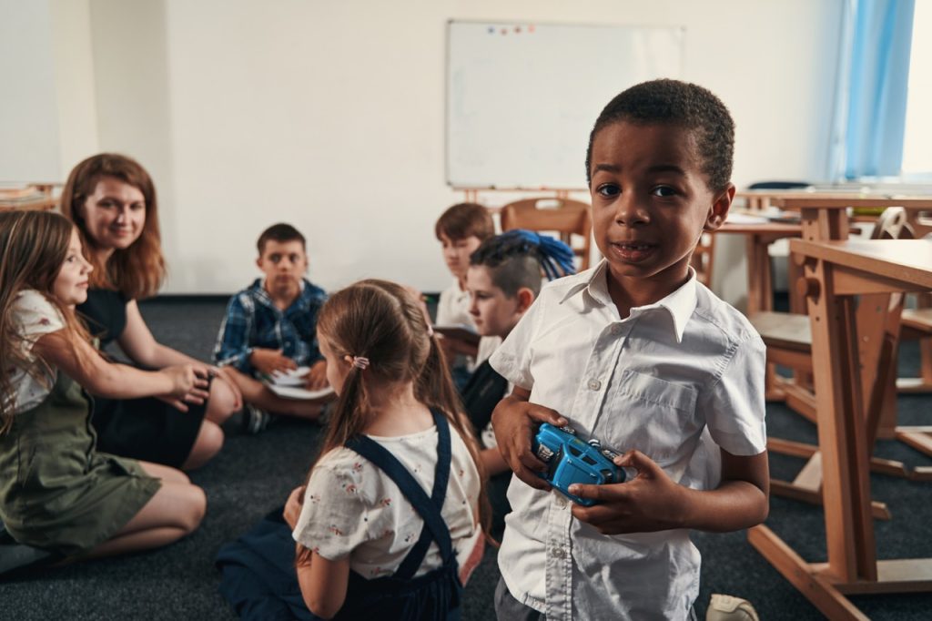 Cute Afro American boy standing with a toy car by the desk. Group of children sitting with a teacher behind him