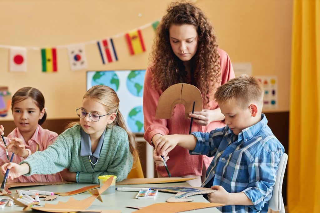 Colorful portrait of kind young teacher assisting children during art and craft class and painting cardboard figures together copy space