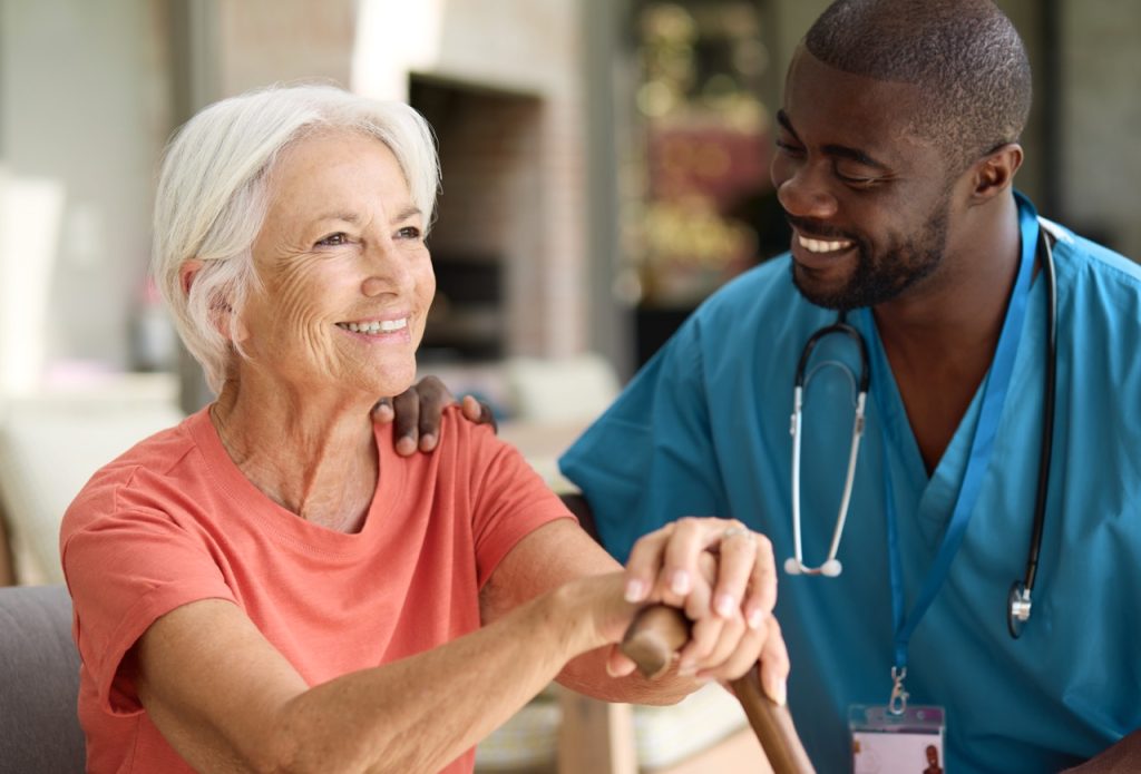 Male Nurse Visiting Senior Woman Using Walking Stick With Mobility Issues At Home