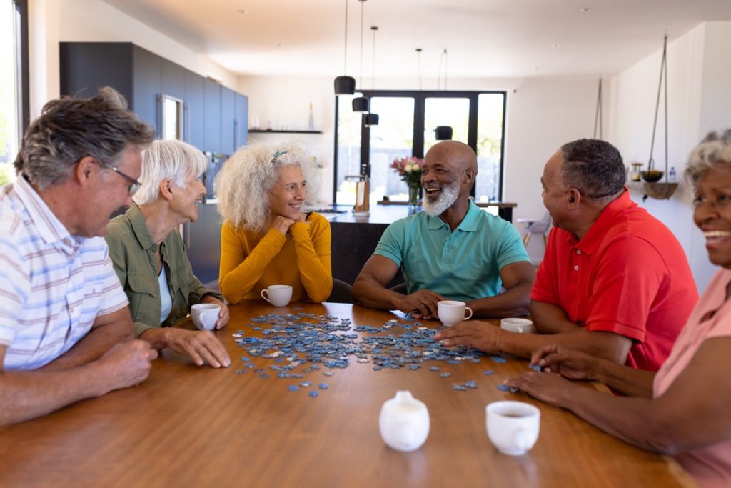 Multiracial seniors friends playing jigsaw puzzle while sitting at wooden table in nursing home. Game, pieces, confusion, brainstorming, unaltered, together, support, assisted living, retirement.