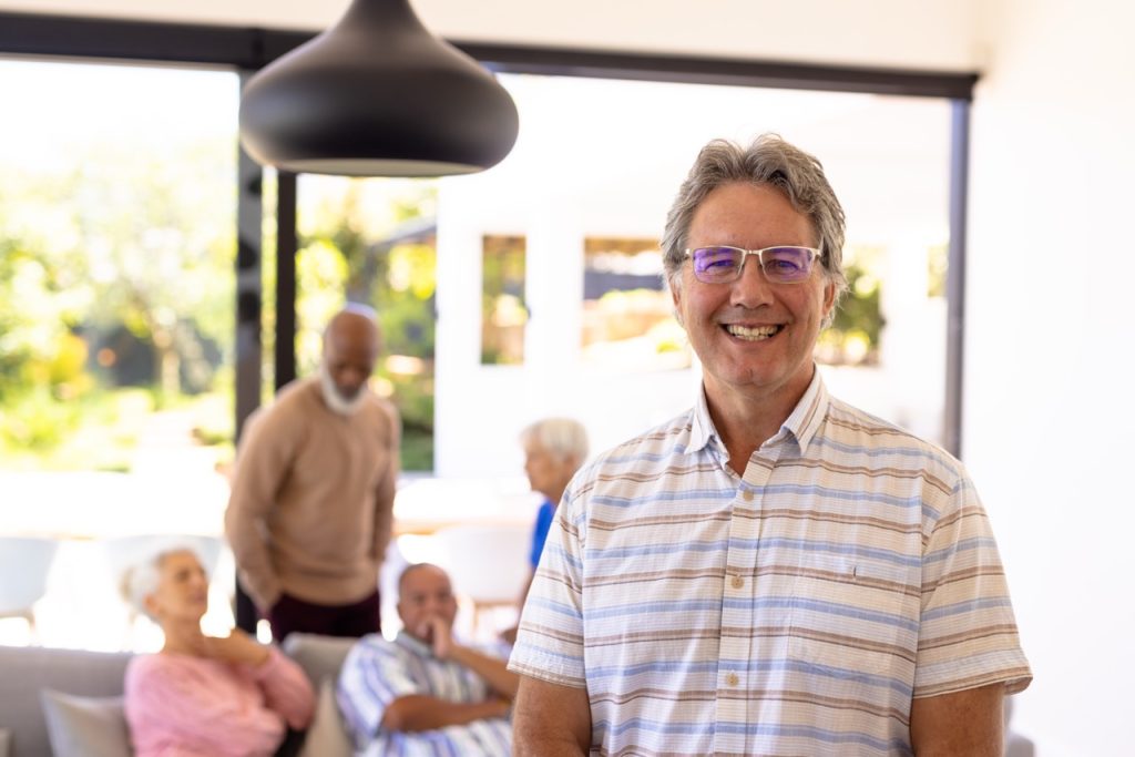 Portrait of smiling caucasian senior man standing with multiracial friends in background. Nursing home, happy, eyeglasses, unaltered, friendship, togetherness, support, assisted living, retirement.