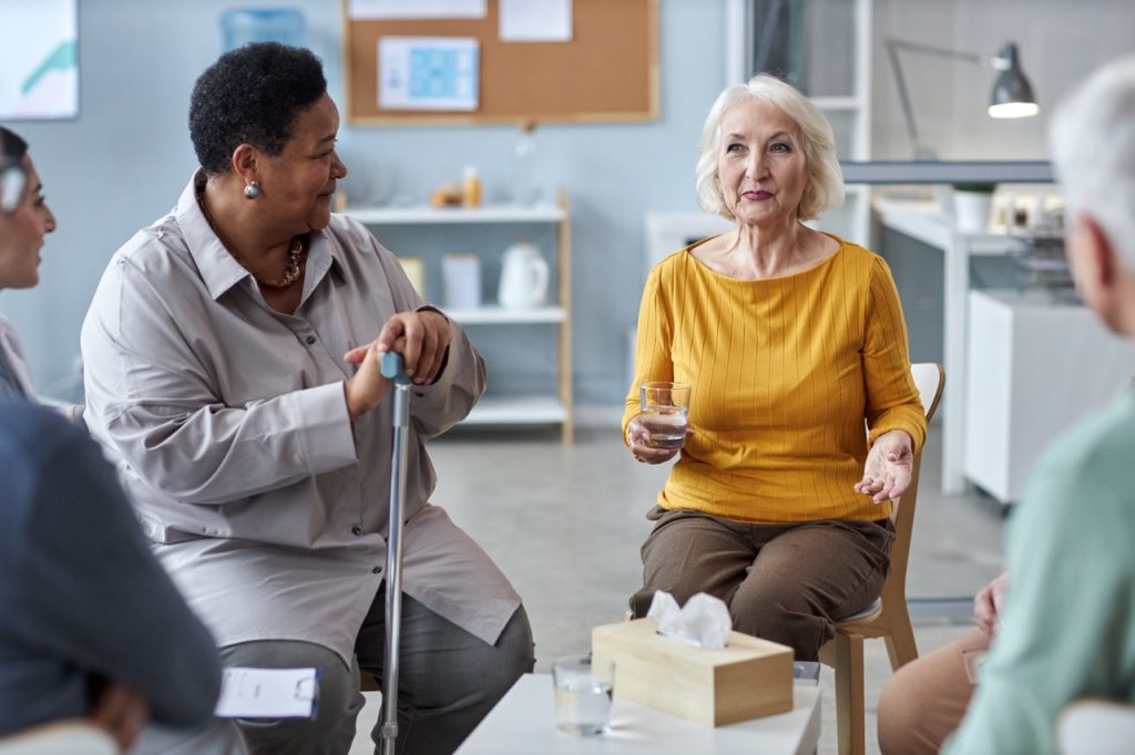 Portrait of two senior women sharing stories while sitting in circle at mental health support group in retirement home