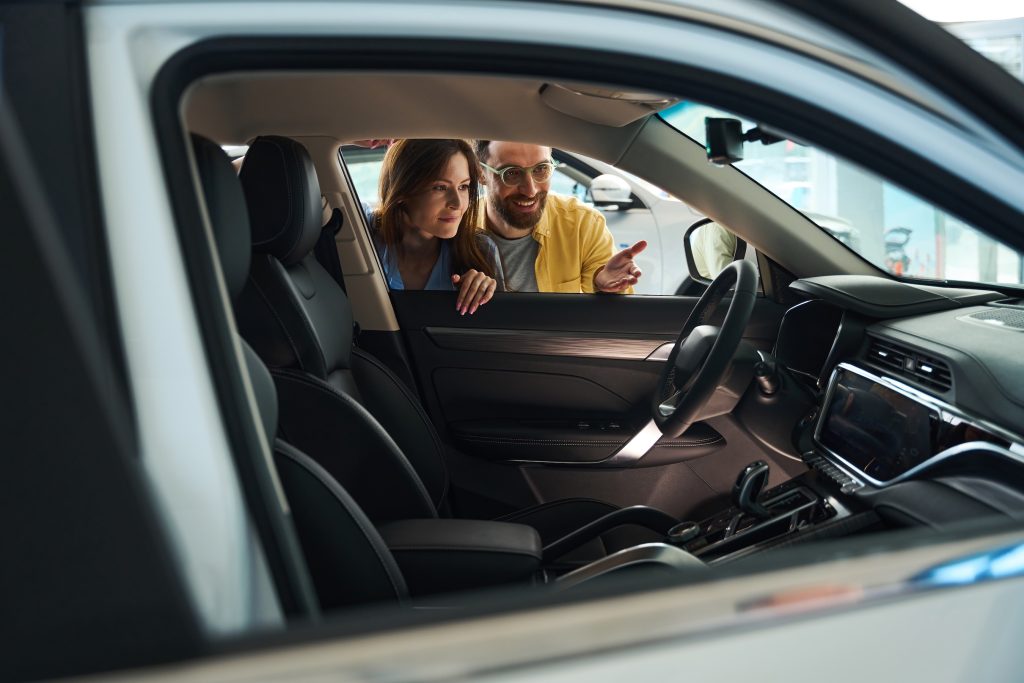 Smiling man in glasses and attractive woman with beautiful manicure looking at interior of auto in car showroom